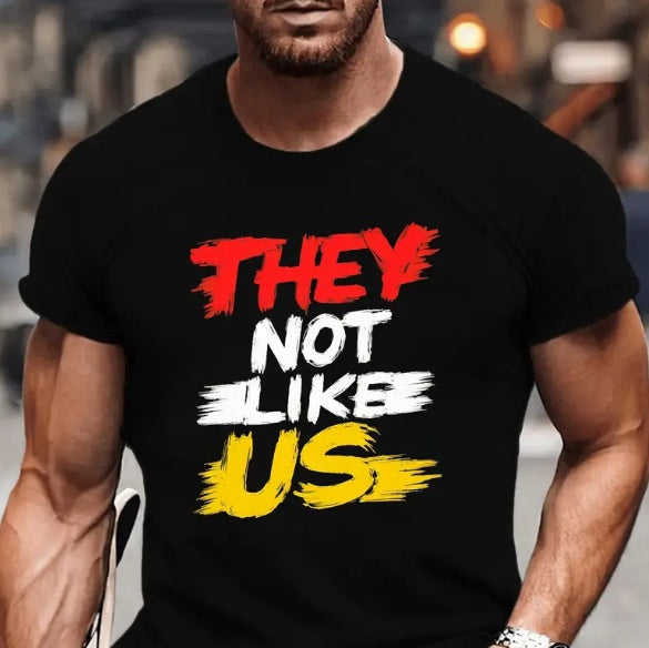 A muscular man wearing a black T-shirt with the phrase "They Not Like Us" in red, white, and yellow brush stroke letters.