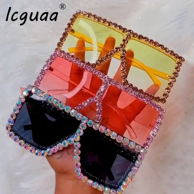 A hand holds three stacked, oversized sunglasses with rhinestone-encrusted frames in yellow, pink, and black.